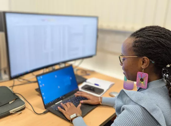 Mary sitting at a computer, working on a spreadsheet. She is wearing a light blue jumper and has large pink earrings. 
