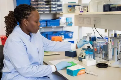 A woman in blue scrubs and gloves sits at a desk, testing liquids with scientific equipment
