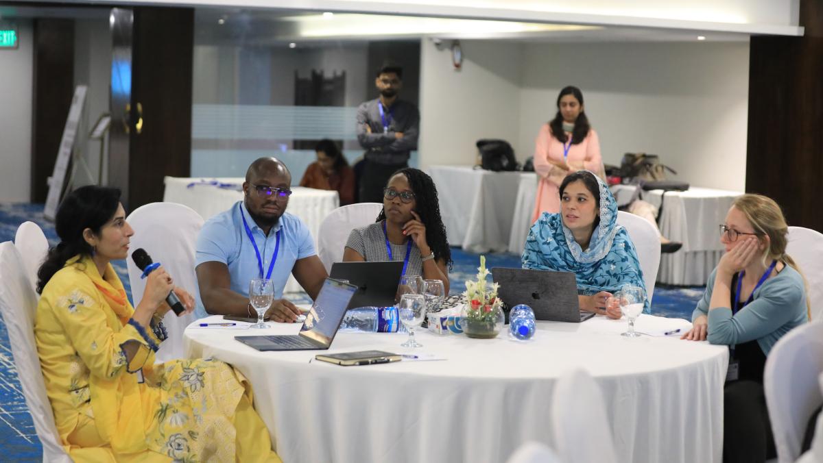 Mary and colleagues sat at a table with a white tablecloth in a conference hall. One of the colleagues is wearing a yellow dress and holding a microphone- the others at the table are watching them. 