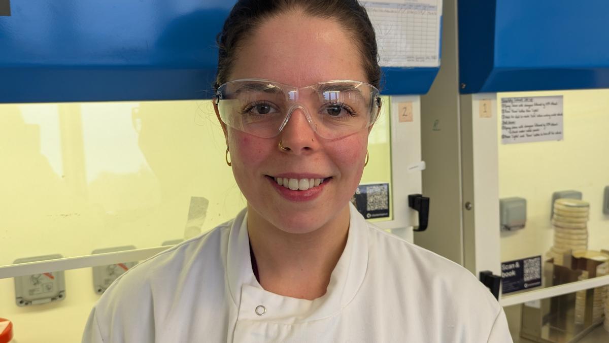 Sarah Langhorne in a lab coat and goggles standing in front of a blue safety cabinet.
