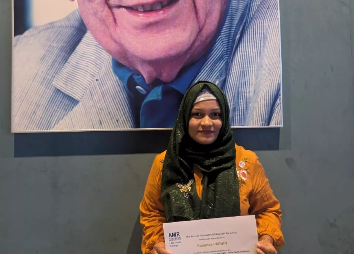 Sahanaz holding her certificate of attendance in front of a portrait of the Merieux foundation founder.